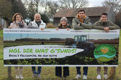 Familie hinter einem großen Werbeschild von Bachl’s Feldgemüse mit dem Slogan „Hol dir was G’sunds!“. Das Schild zeigt ein Gemüsefeld mit Traktor und informiert über die Ab-Hof-Hütte in Streitdorf.